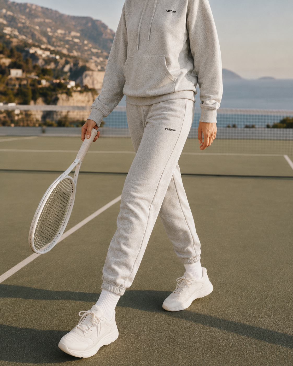 Pantalon de survêtement femme Kardaia gris en coton biologique, photographié en mouvement sur un court de tennis. Sa coupe décontractée et ses chevilles élastiquées sont associées au sweat à capuche gris coordonné.